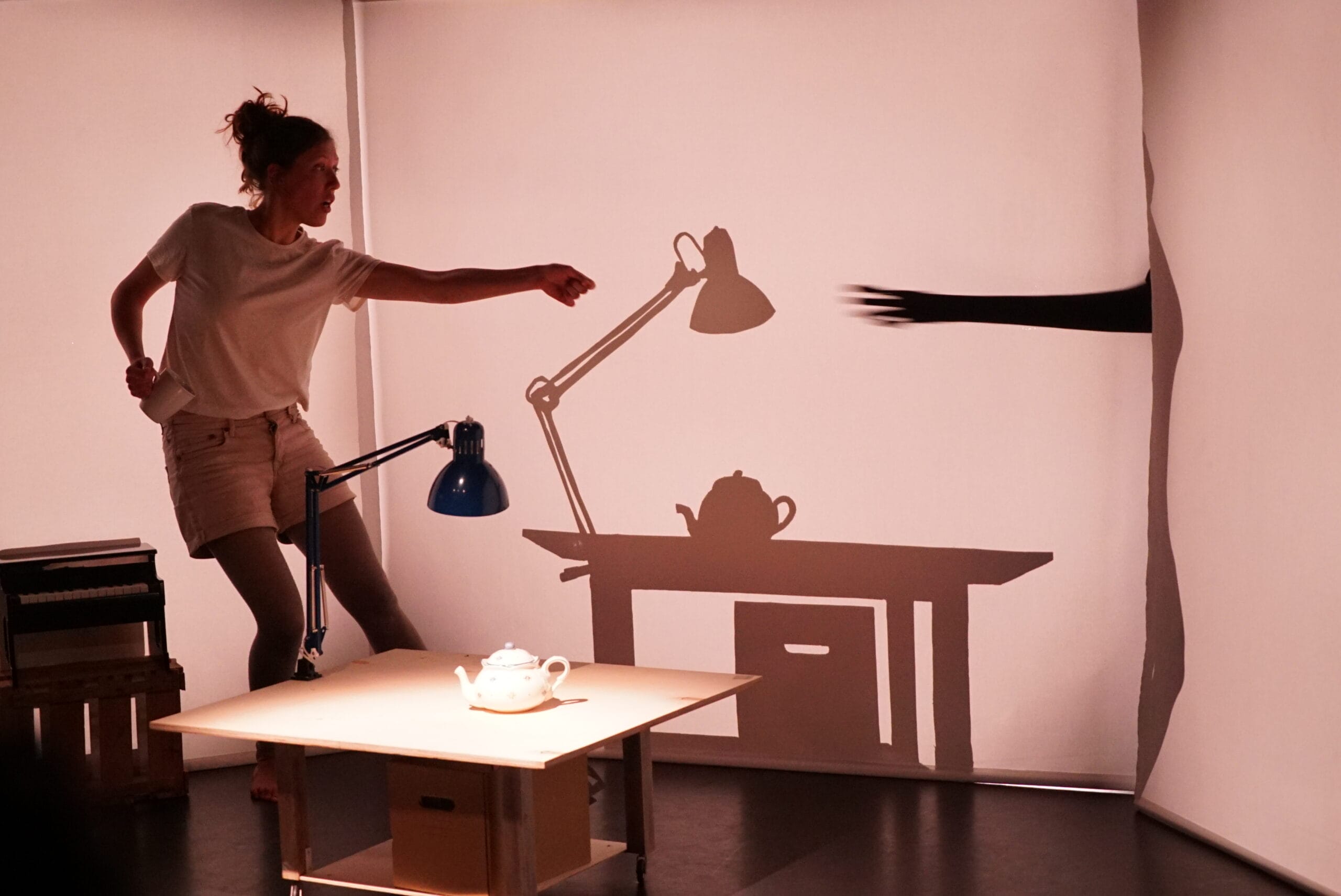 A woman stands next to a desk, reaching toward a lamp. The scene's shadow shows a hand reaching through a tear in the backdrop toward the lamp and teapot.