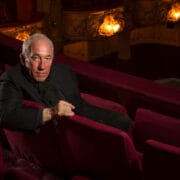 Simon Callow in a dark suit sits alone in a theatre's red velvet seats, with ornate balconies and warm lights visible in the background.