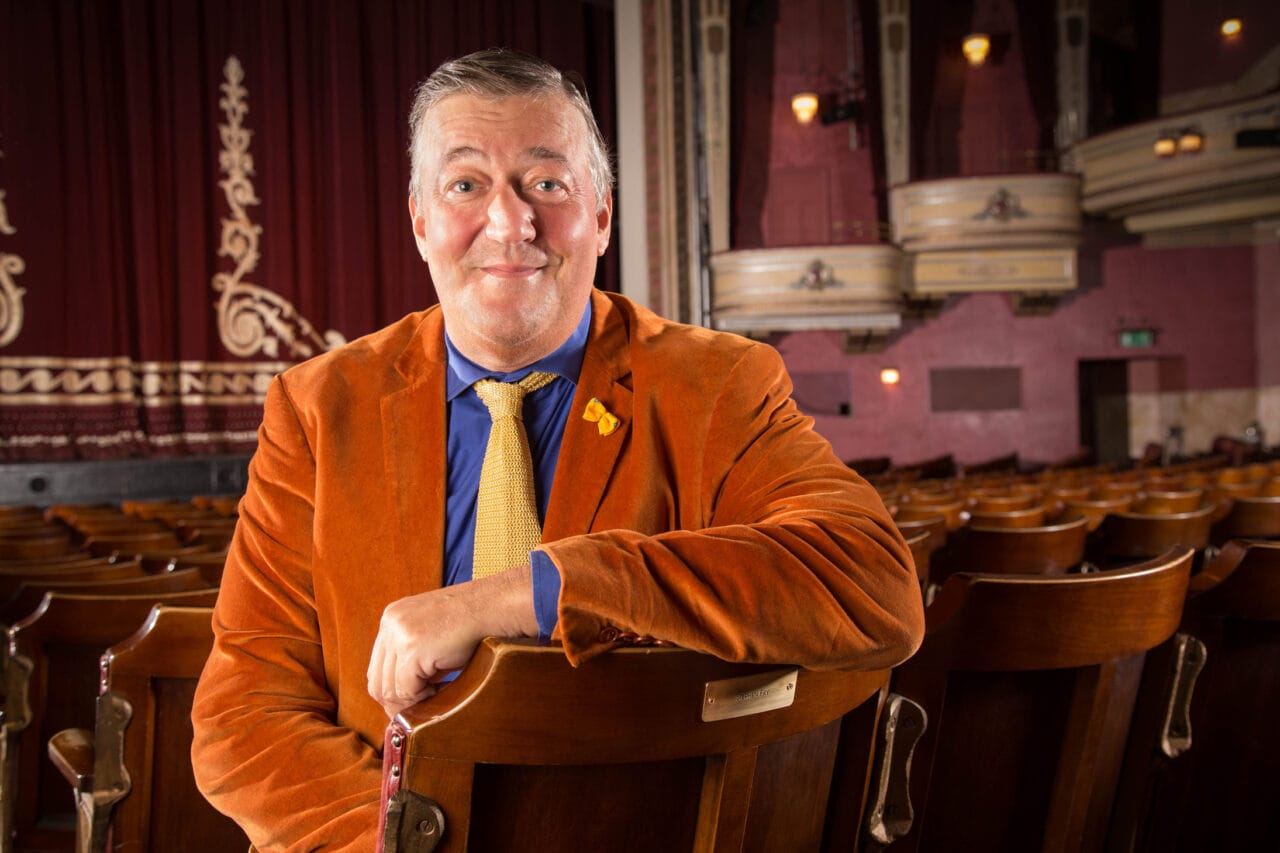 Stephen Fry in an orange jacket and yellow tie sits on a theatre seat, facing the camera, with an empty theatre and stage in the background.