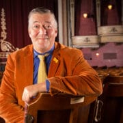 Stephen Fry in an orange jacket and yellow tie sits on a theatre seat, facing the camera, with an empty theatre and stage in the background.