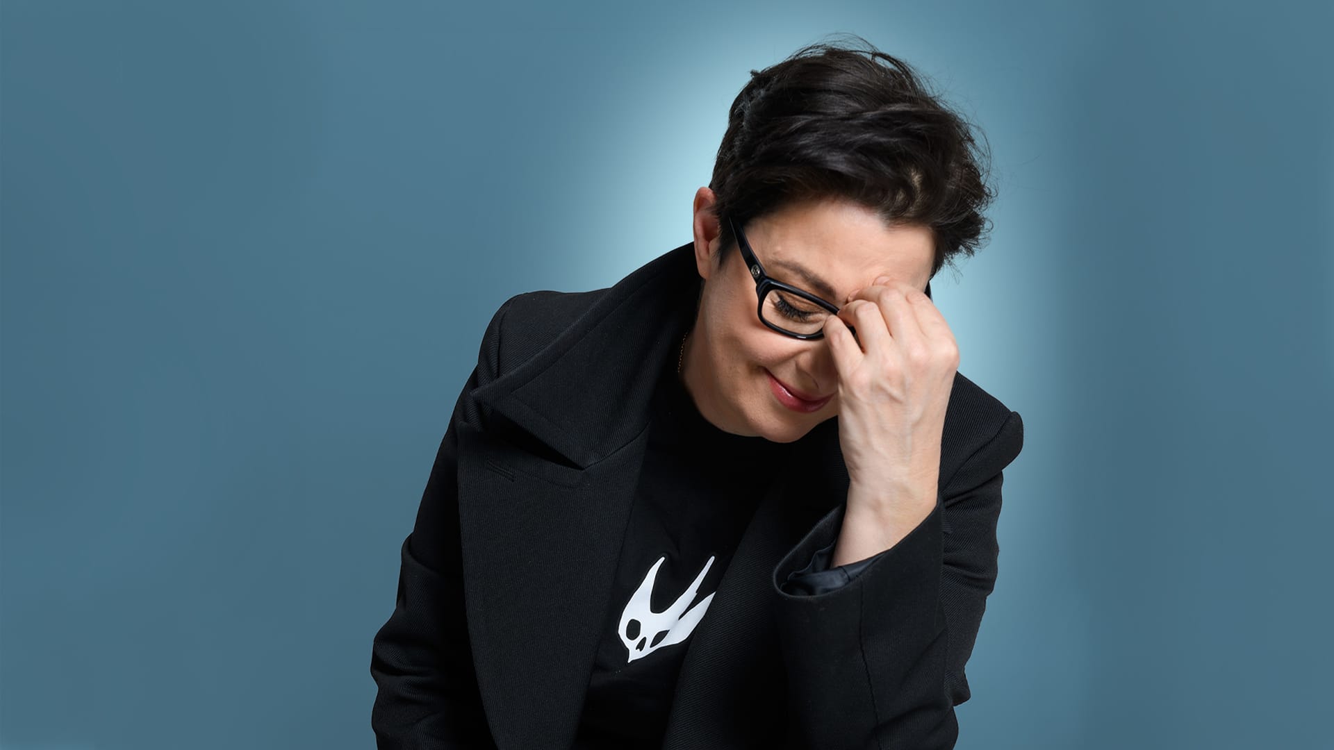 Person with short dark hair and glasses sits against a blue background, smiling with eyes closed and hand covering part of face.