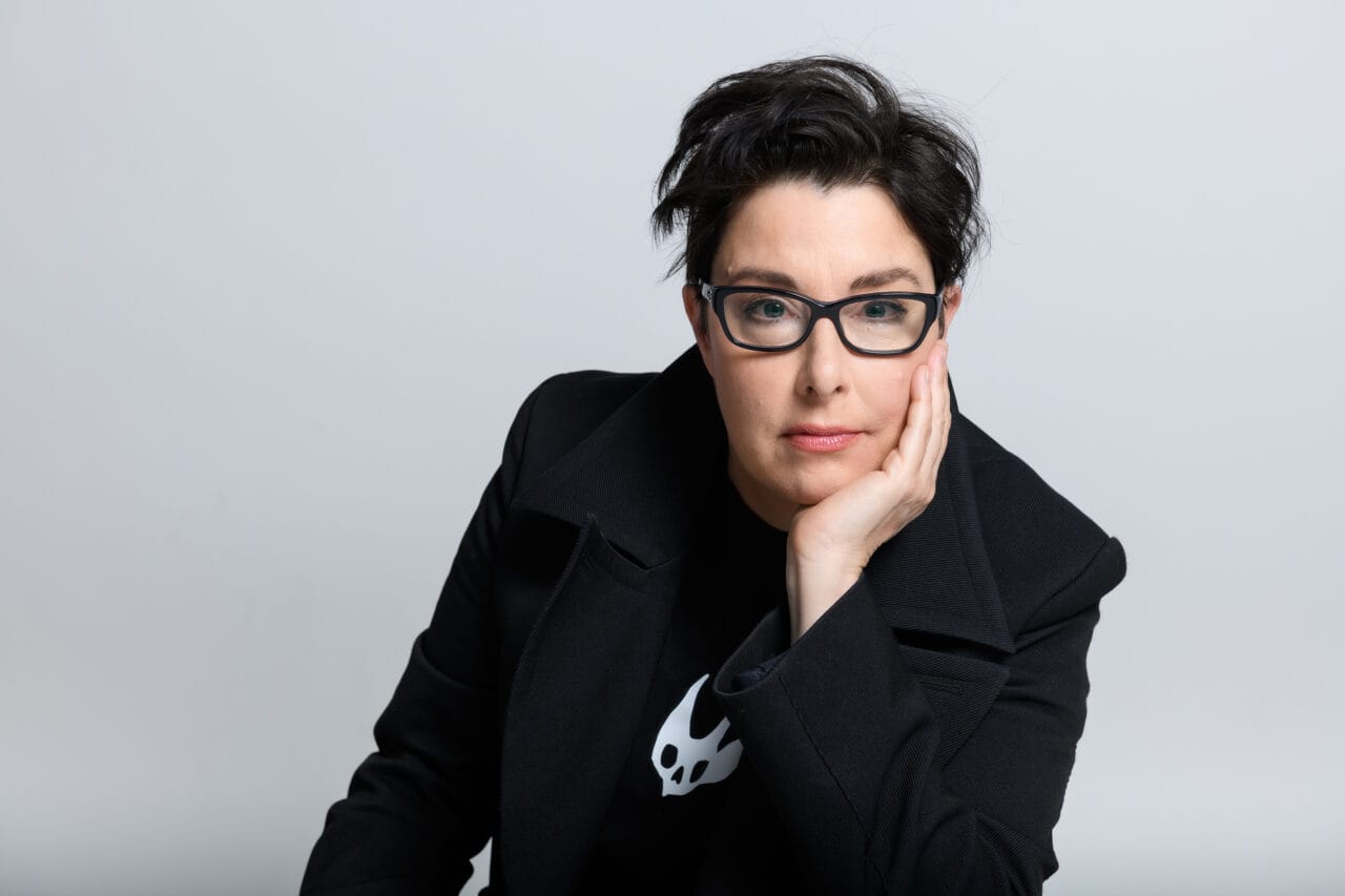Person with short dark hair and glasses, wearing a black coat and resting their head on one hand, looks directly at the camera against a plain light background.