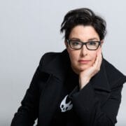 Person with short dark hair and glasses, wearing a black coat and resting their head on one hand, looks directly at the camera against a plain light background.