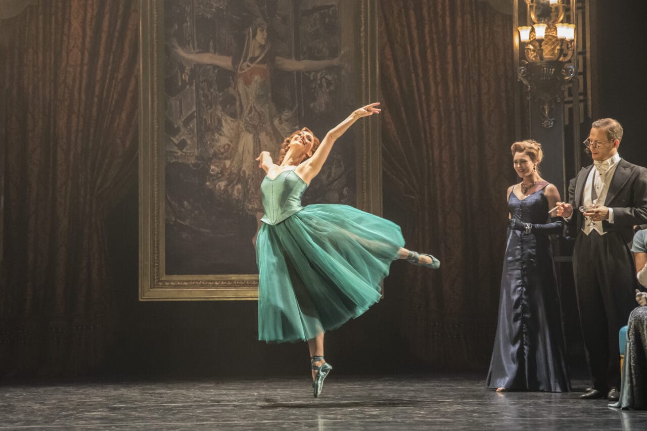 A ballerina in a teal dress performs on stage in front of a large painting, while three formally dressed people watch from the side.
