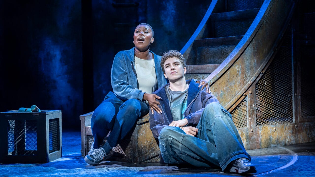 Two actors sit at the base of a large metal structure on stage, one singing with an expressive face while the other looks thoughtful—both dressed in casual clothing, capturing the spirit of The Lightning Thief: The Percy Jackson Musical.