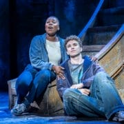 Two actors sit at the base of a large metal structure on stage, one singing with an expressive face while the other looks thoughtful—both dressed in casual clothing, capturing the spirit of The Lightning Thief: The Percy Jackson Musical.