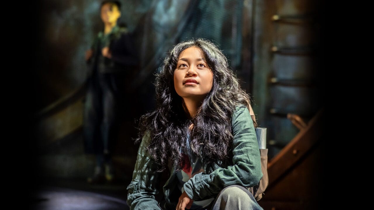 A young woman with long, wavy hair sits and gazes upward thoughtfully in a dimly lit setting, reminiscent of a scene from The Lightning Thief: The Percy Jackson Musical, with another person blurred in the background.