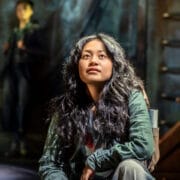 A young woman with long, wavy hair sits and gazes upward thoughtfully in a dimly lit setting, reminiscent of a scene from The Lightning Thief: The Percy Jackson Musical, with another person blurred in the background.