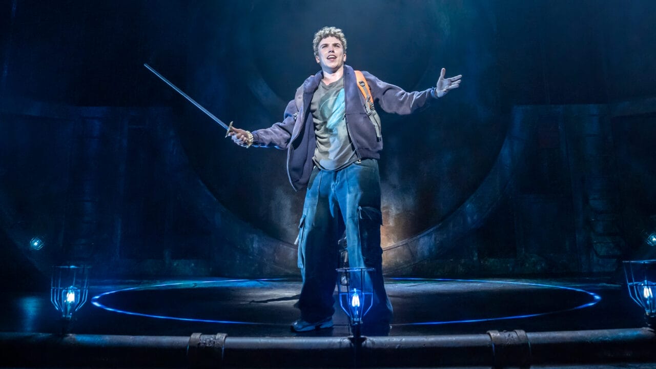 A young man stands on stage holding a sword, dressed in a jacket and cargo pants, with blue lighting and circular stage elements behind him—a scene reminiscent of the Percy Jackson Musical, inspired by The Lightning Thief.