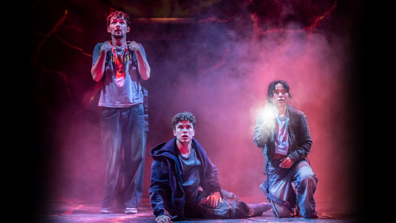 Three young people in casual, worn clothes stand and kneel on a smoky stage, with one holding a glowing light, evoking a dramatic scene from the Percy Jackson musical, The Lightning Thief.