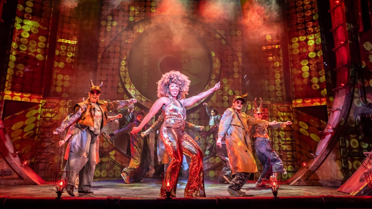 A performer in a gold sequin outfit dances center stage with backup dancers in costumes, under dramatic lighting and colorful stage effects inspired by The Lightning Thief: The Percy Jackson musical.