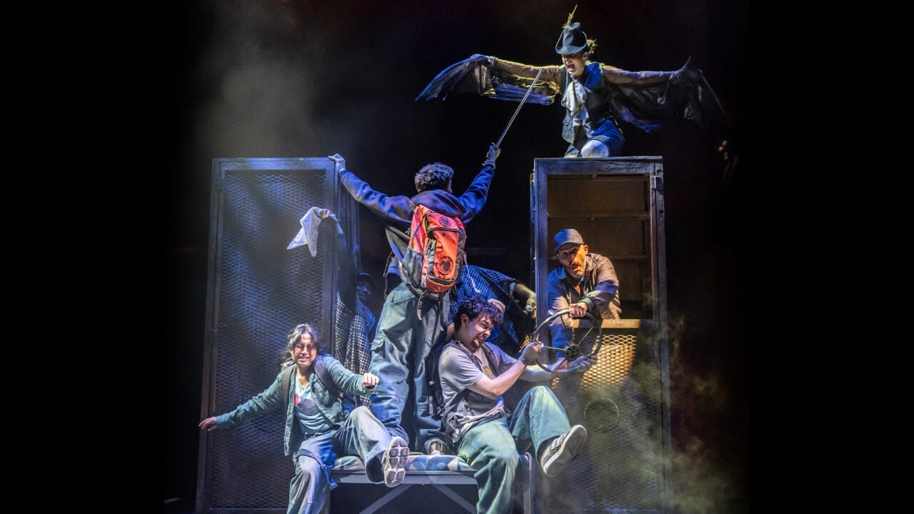 Five actors perform on stage with dramatic lighting in this electrifying Percy Jackson Musical scene; one, wearing wings, stands above the others as they energetically interact with props and each other.