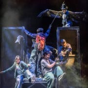 Five actors perform on stage with dramatic lighting in this electrifying Percy Jackson Musical scene; one, wearing wings, stands above the others as they energetically interact with props and each other.