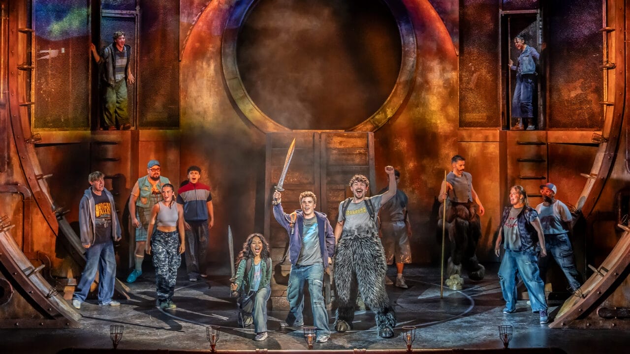 A group of actors performs energetically on stage with dramatic lighting and costumes, capturing the excitement of The Lightning Thief: The Percy Jackson Musical, featuring one person holding a sword and others raising their fists in the air.