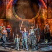 A group of actors performs energetically on stage with dramatic lighting and costumes, capturing the excitement of The Lightning Thief: The Percy Jackson Musical, featuring one person holding a sword and others raising their fists in the air.