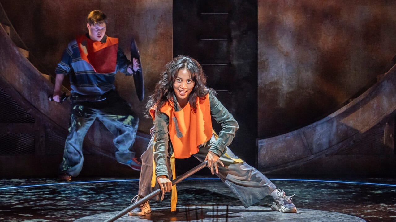 Two actors on stage, dressed in casual clothing and orange armor vests, perform a dramatic scene inspired by The Lightning Thief: The Percy Jackson Musical, with props against a dimly lit industrial backdrop.