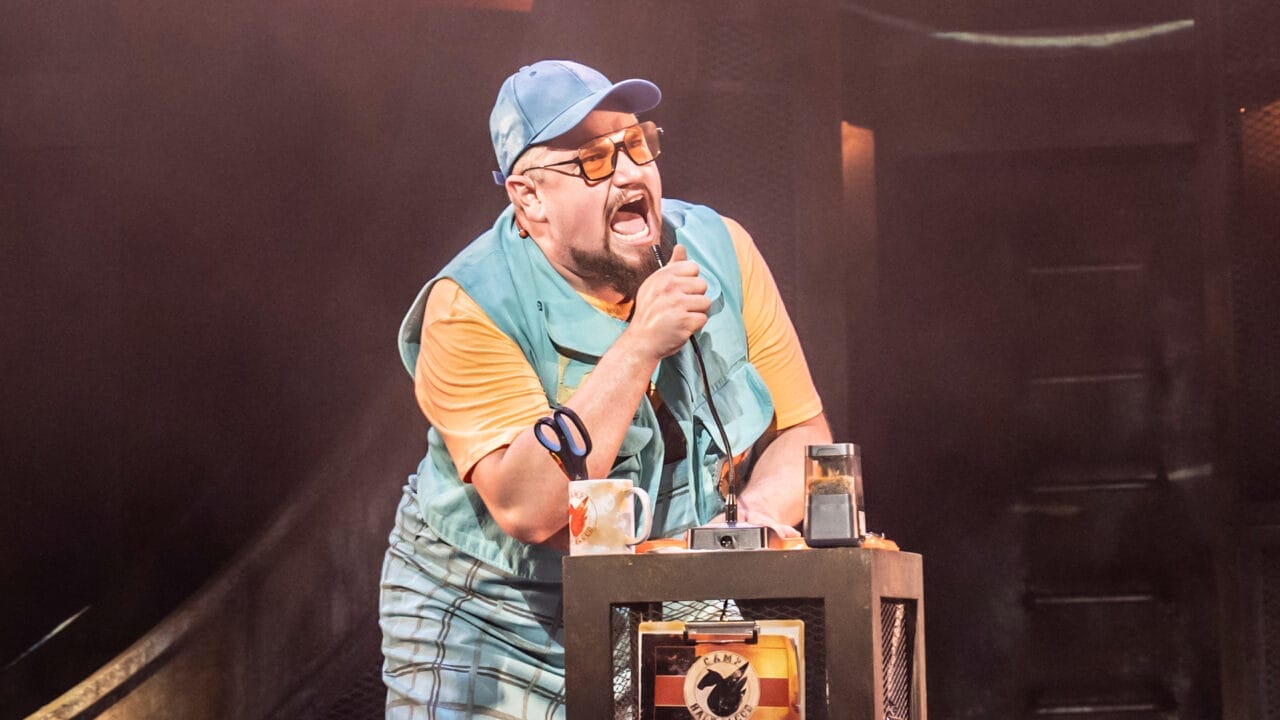 A man wearing glasses, a blue cap, and a vest passionately speaks or sings into a microphone behind a small desk—evoking the energy of The Lightning Thief: The Percy Jackson Musical.