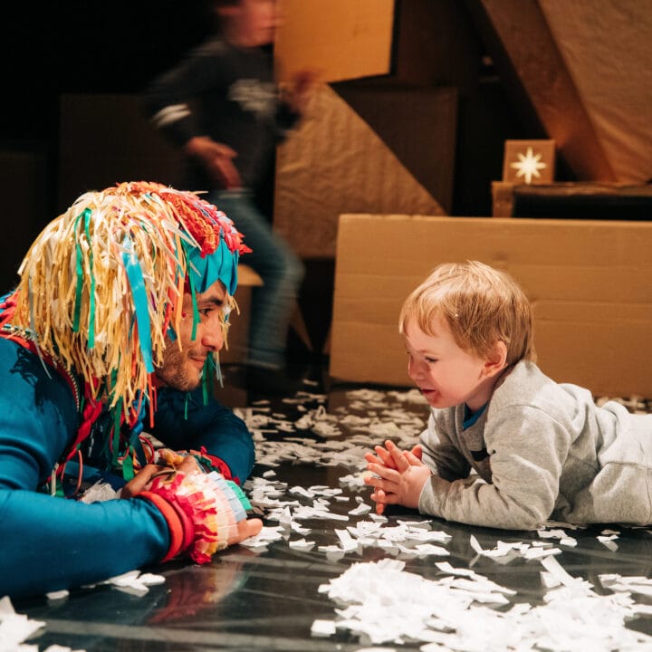 An adult in a colorful costume lies on the floor facing a smiling child, both surrounded by pieces of white paper with cardboard boxes in the background.