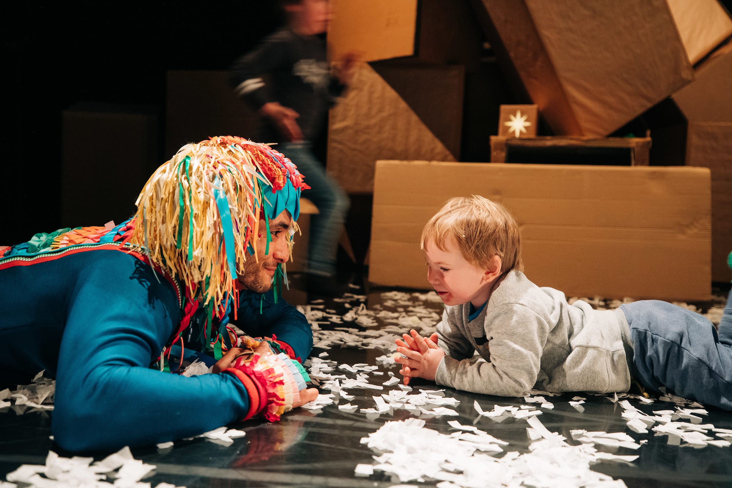 An adult in a colorful costume lies on the floor facing a smiling child, both surrounded by pieces of white paper with cardboard boxes in the background.