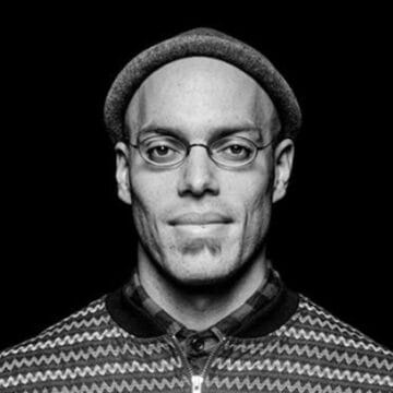 A man wearing glasses and a hat faces forward, looking at the camera, against a plain black background.