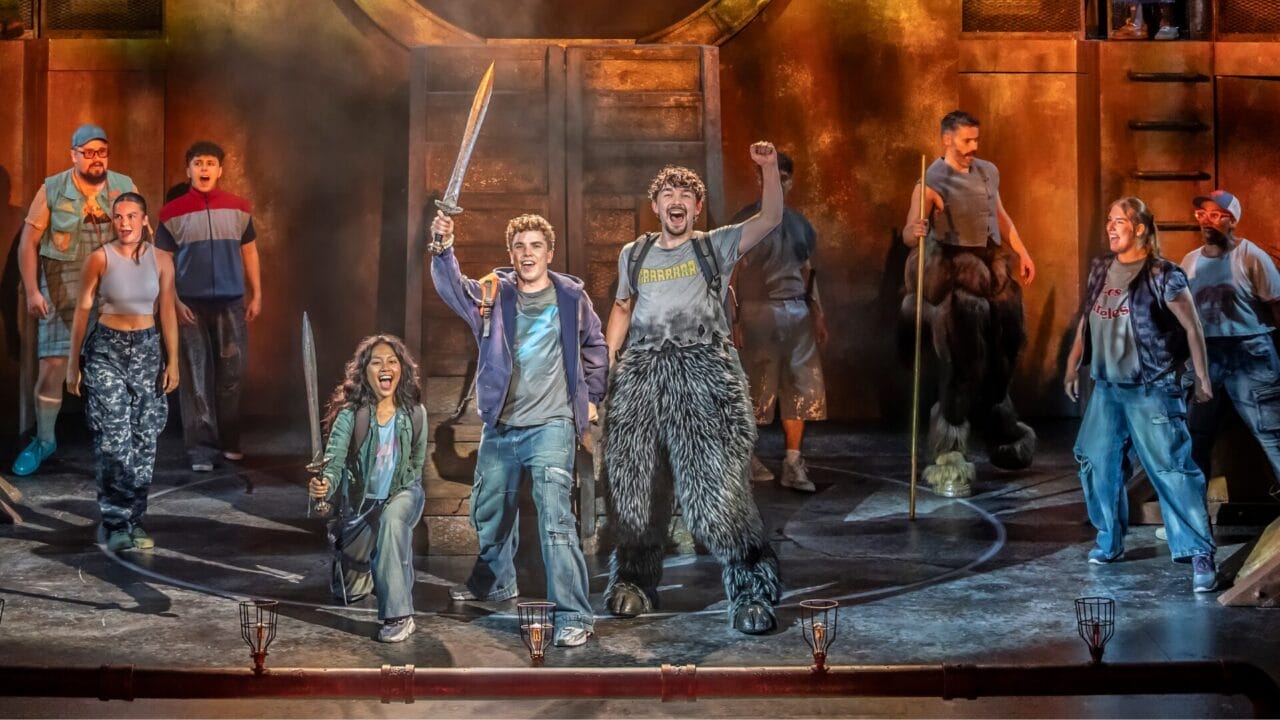 Three young actors in costume stand on stage, swords in hand and fists raised triumphantly, bringing the world of The Lightning Thief: The Percy Jackson Musical to life against a dramatic wooden set and moody lighting.