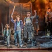 Three young actors in costume stand on stage, swords in hand and fists raised triumphantly, bringing the world of The Lightning Thief: The Percy Jackson Musical to life against a dramatic wooden set and moody lighting.