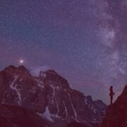 Two rock climbers in the Red Programme scale a cliff at night beneath a star-filled sky, with a large mountain rising in the background.
