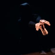 A person in a dark hoodie extends their arms forward, holding several small mannequin hands against a black background.