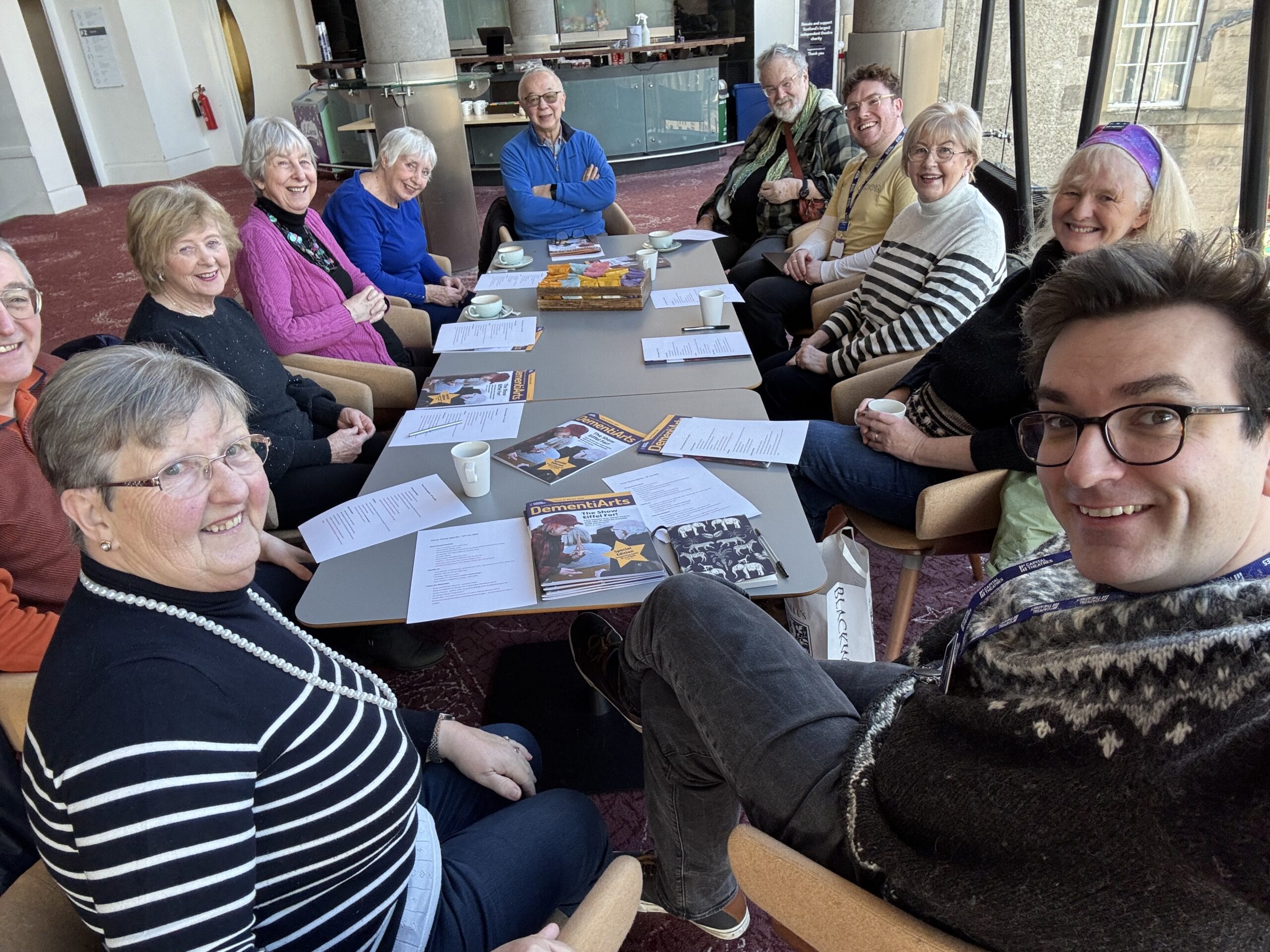 A group of people seated around a table, smiling. Papers and magazines are on the table. A person in the foreground takes a selfie, capturing the group meeting.