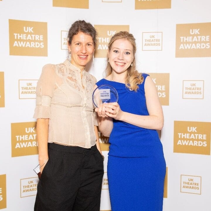 Two women pose for a photo at the UK Theatre Awards, one holding an award. They stand against a backdrop with the event's logo.