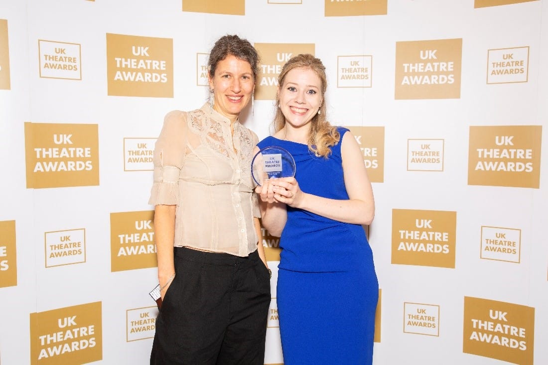 Two women pose for a photo at the UK Theatre Awards, one holding an award. They stand against a backdrop with the event's logo.