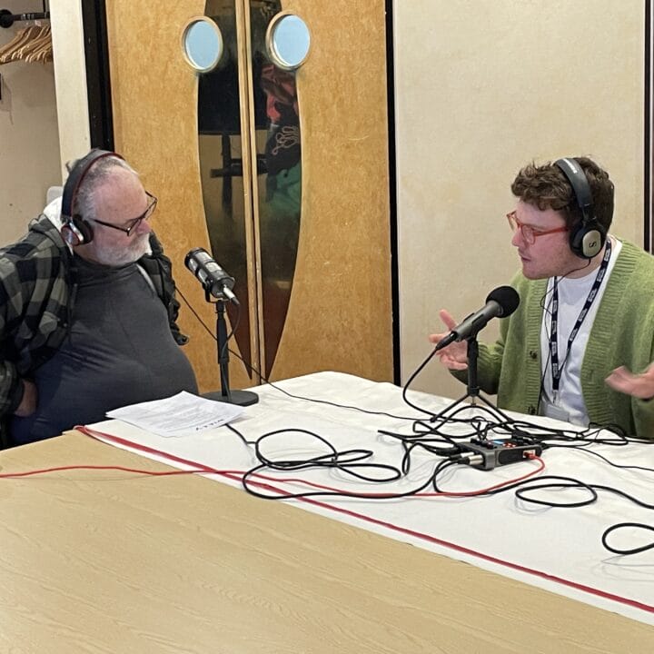 Two people wearing headphones sit at a table with microphones, engaged in conversation or podcast recording in an indoor setting.