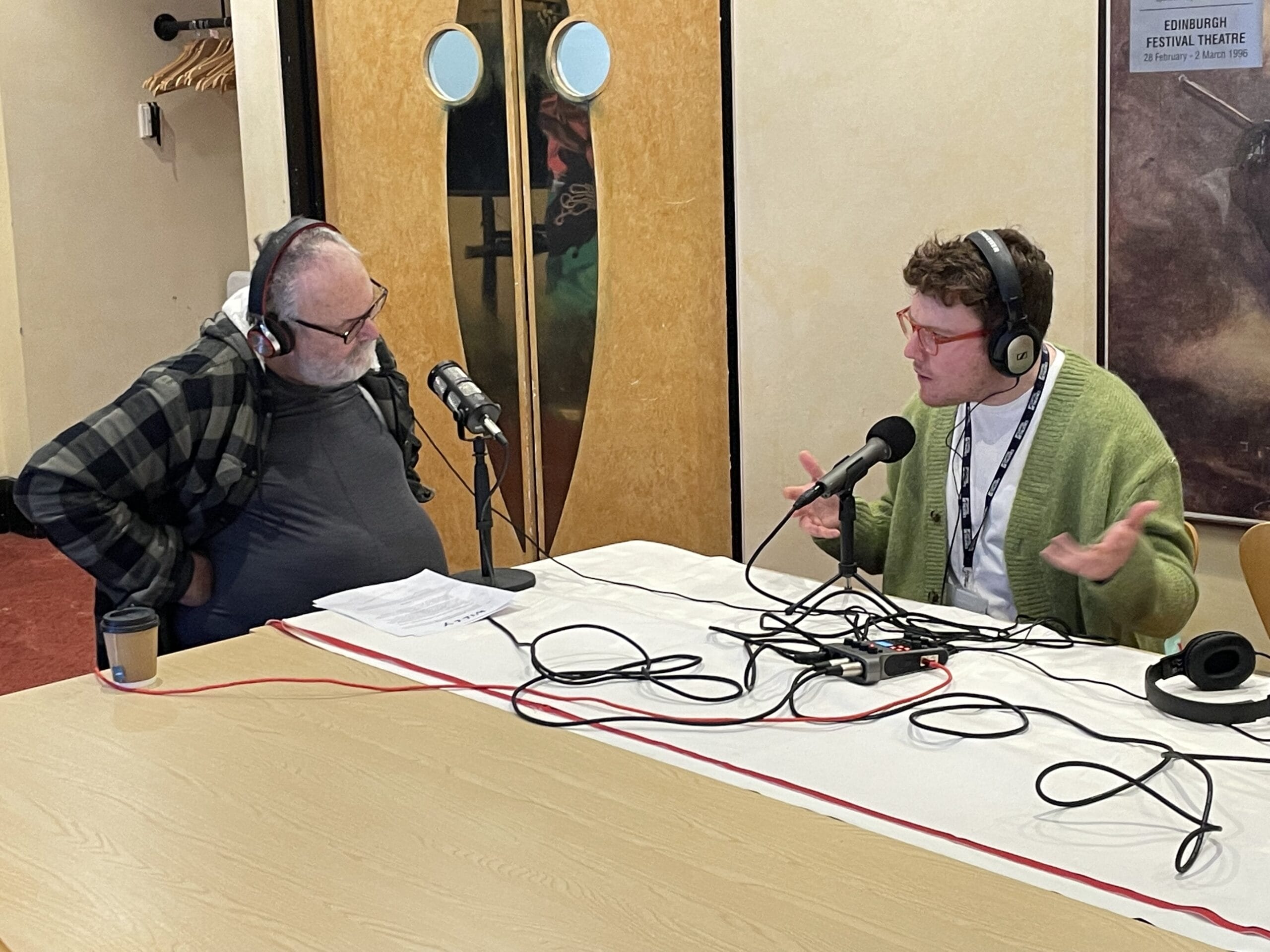 Two people wearing headphones sit at a table with microphones, engaged in conversation or podcast recording in an indoor setting.