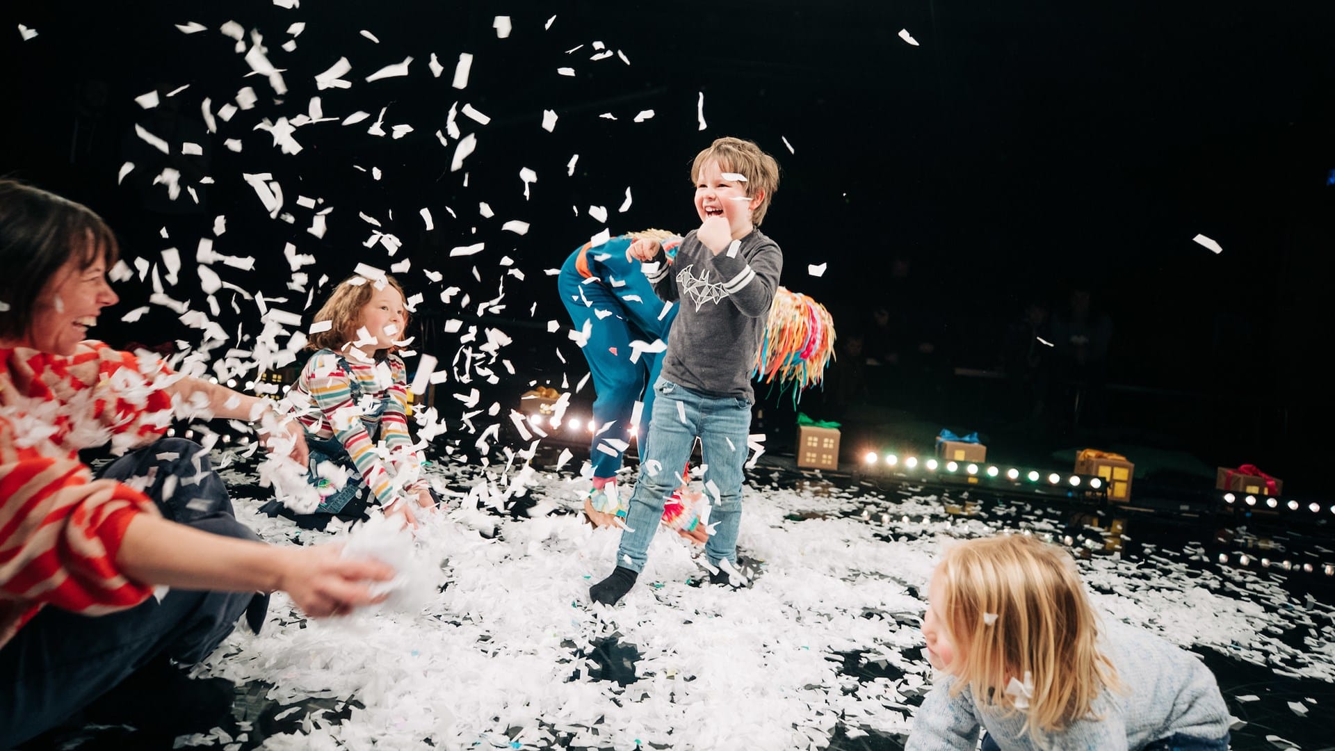 Children playing in artificial snow on a dark stage, laughing and playing while an adult watches them. Multicolored lights and miniature house props are in the background.
