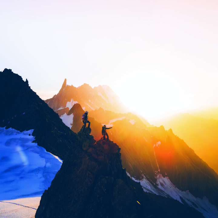 Two hikers stand on a rocky mountain ridge at sunrise, with snow-covered slopes and distant peaks illuminated by orange sunlight.
