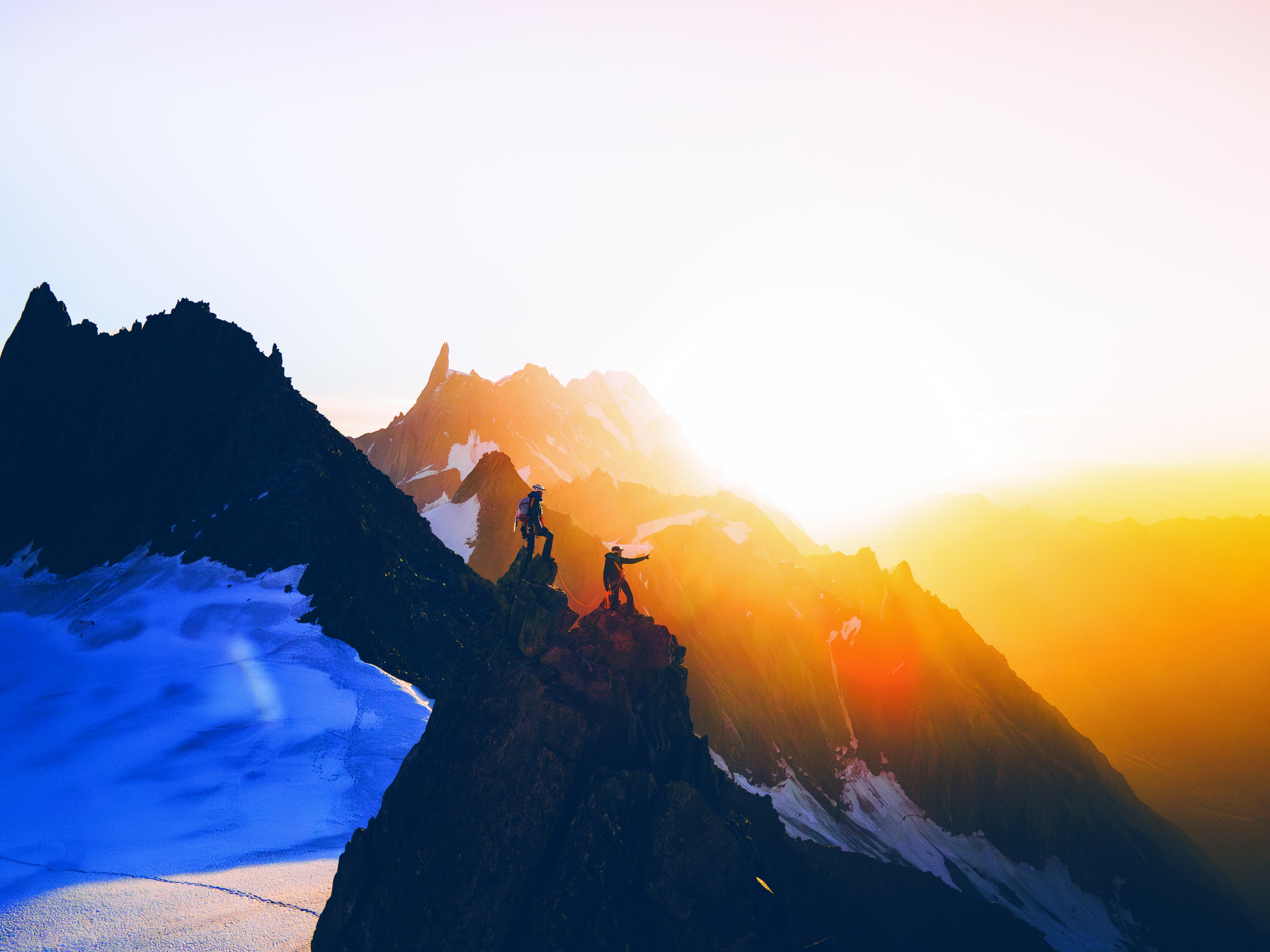 Two hikers stand on a rocky mountain ridge at sunrise, with snow-covered slopes and distant peaks illuminated by orange sunlight.