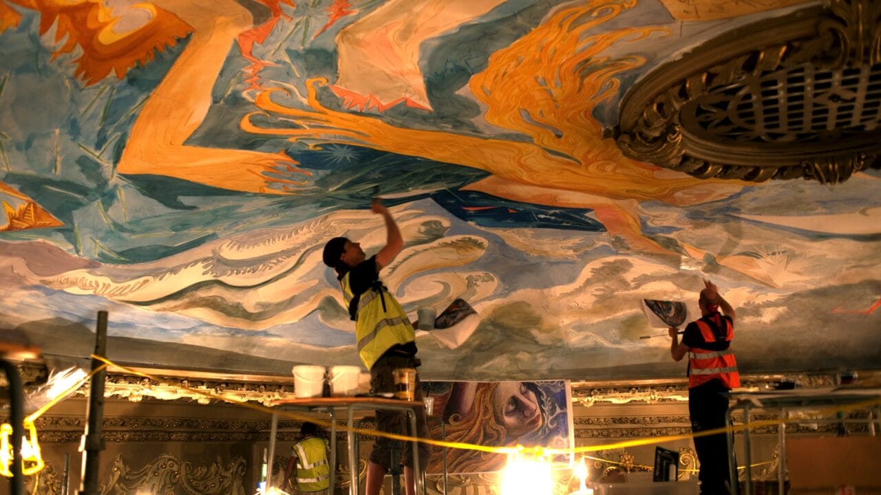 Two workers in safety vests restore the King's Theatre's colorful, ornate ceiling mural, using tools and scaffolding under bright lighting.
