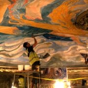 Two workers in safety vests restore the King's Theatre's colorful, ornate ceiling mural, using tools and scaffolding under bright lighting.
