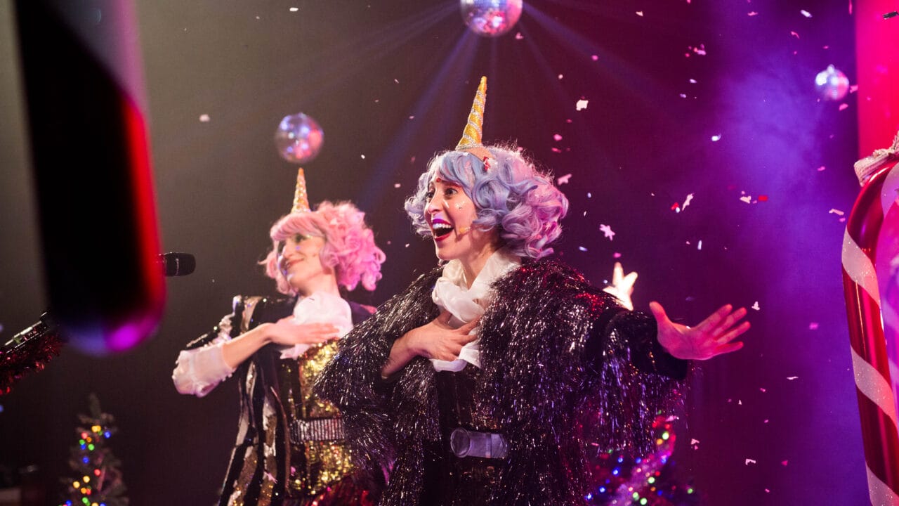 Two performers with colorful wigs and unicorn horns sing on stage amid falling confetti, disco balls, and festive lighting.