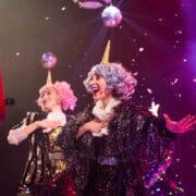 Two performers with colorful wigs and unicorn horns sing on stage amid falling confetti, disco balls, and festive lighting.
