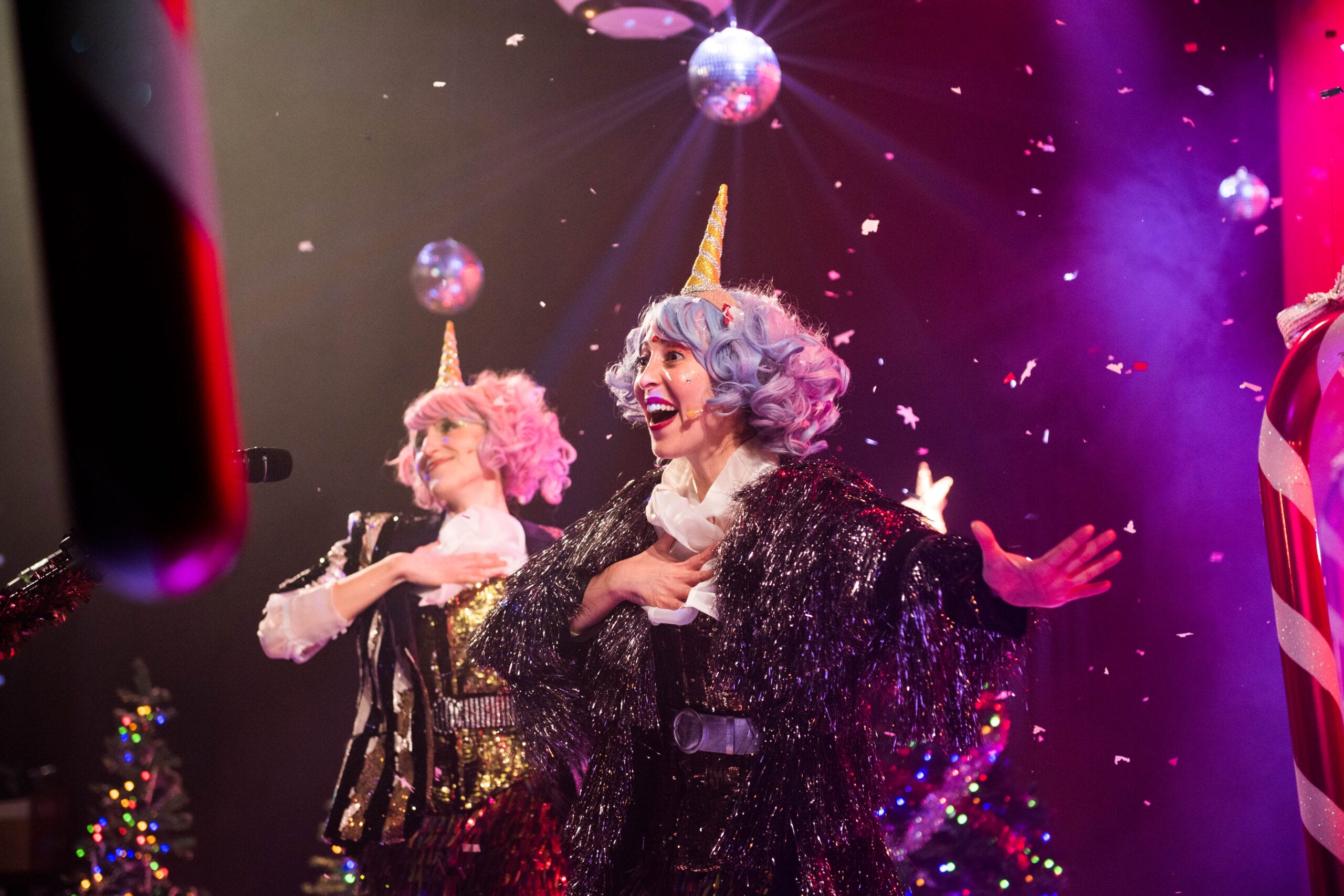 Two performers with colorful wigs and unicorn horns sing on stage amid falling confetti, disco balls, and festive lighting.
