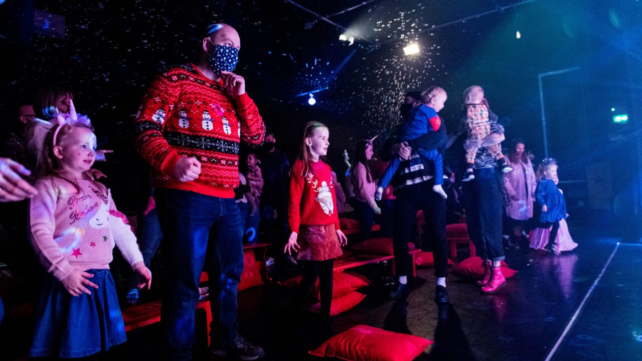A group of children and adults, some in festive jumpers, stand in a dark room with falling artificial snow and stage lighting.