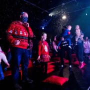 A group of children and adults, some in festive jumpers, stand in a dark room with falling artificial snow and stage lighting.