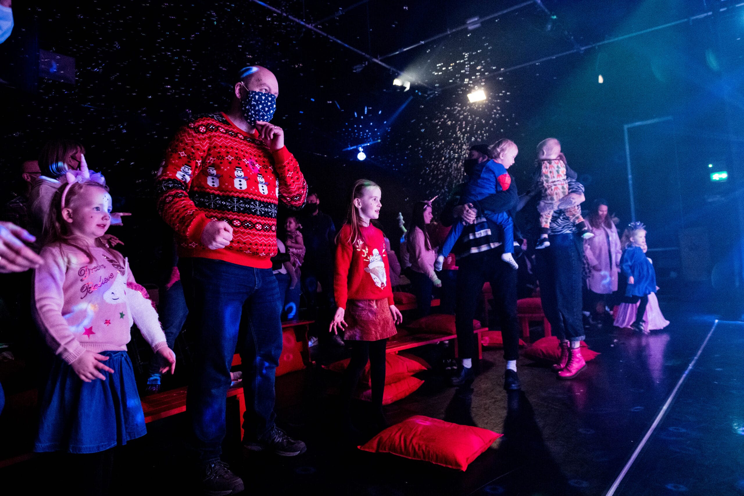 A group of children and adults, some in festive jumpers, stand in a dark room with falling artificial snow and stage lighting.