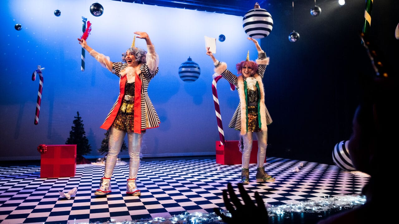 Two performers in colorful, clown-like costumes stand on a checkered stage decorated with oversized ornaments and candy canes, waving to an audience with hands visible in the foreground.