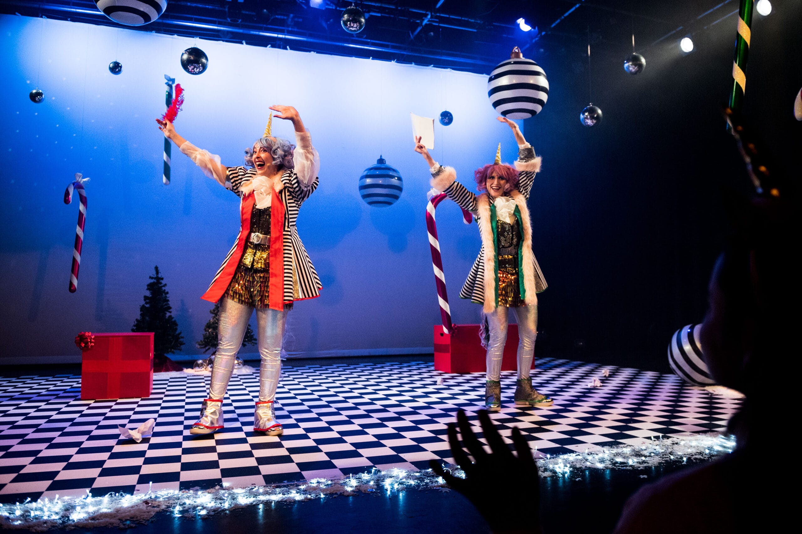 Two performers in colorful, clown-like costumes stand on a checkered stage decorated with oversized ornaments and candy canes, waving to an audience with hands visible in the foreground.