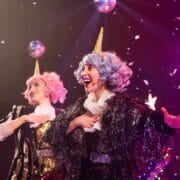Two performers with pastel wigs and unicorn horn headbands sing on stage under confetti and disco balls, dressed in shiny and glittery costumes with dramatic lighting.