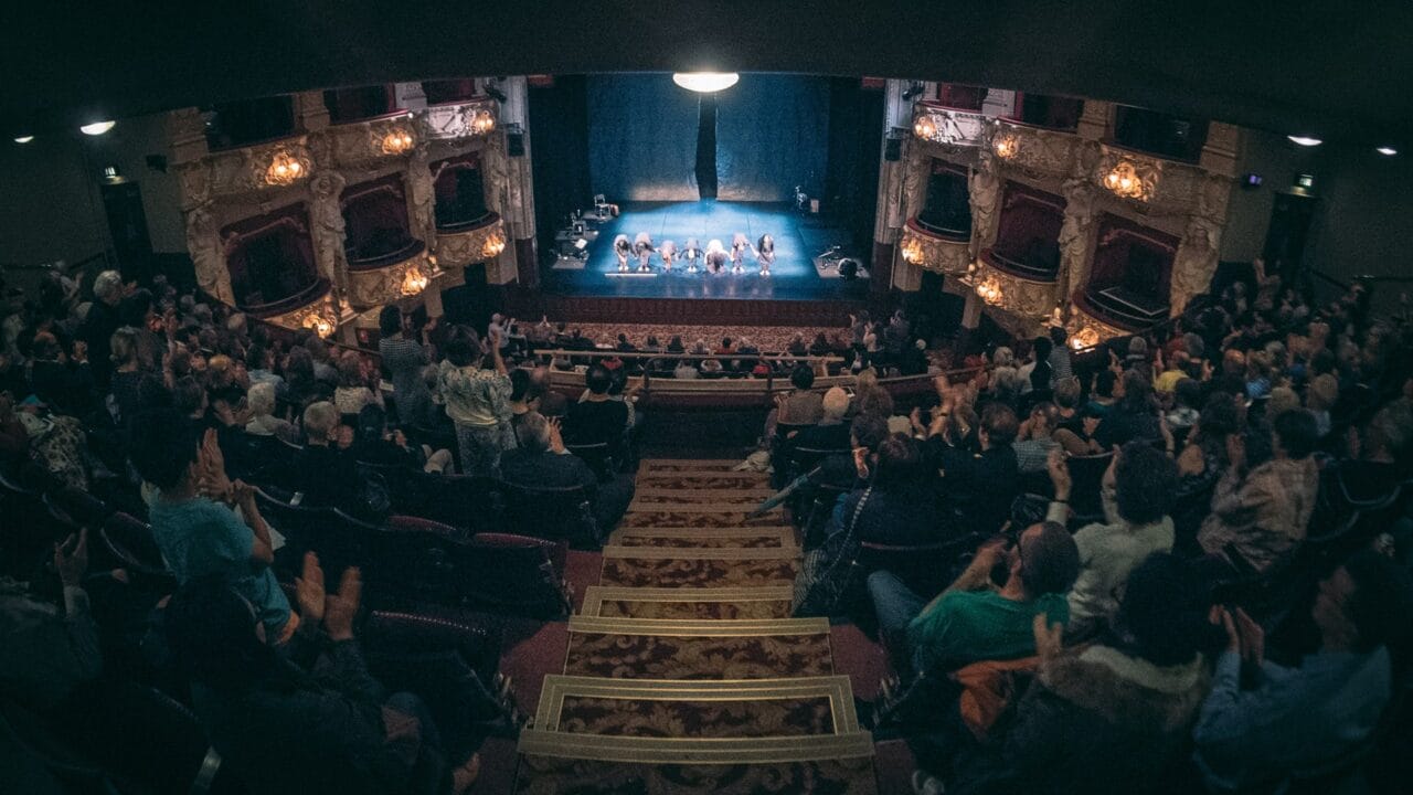 Audience in a theater gives a standing ovation as a group of performers bow on stage under dim lighting, with ornate balconies visible on both sides.