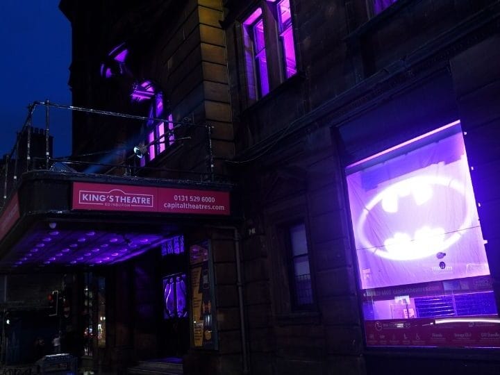 The exterior of King's Theatre at night is illuminated with purple lights, and a large Batman symbol is projected onto a window.