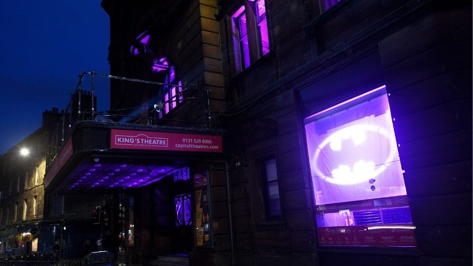 The exterior of King's Theatre at night is illuminated with purple lights, and a large Batman symbol is projected onto a window.